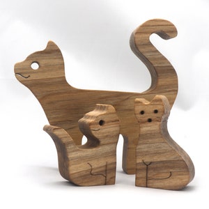 A freestanding wooden puzzle cut in the shape of a large cat standing on four legs. Two smaller kitten figures nest on the large cat’s back and interlock as puzzle pieces. Simple drilled holes form the eyes and nose on each cat, with a shallow cut line for the mouth on the large cat. The completed puzzle stands upright on a plain white surface. Handmade by Odin’s Toy Factory In Tallahassee, Florida, USA.