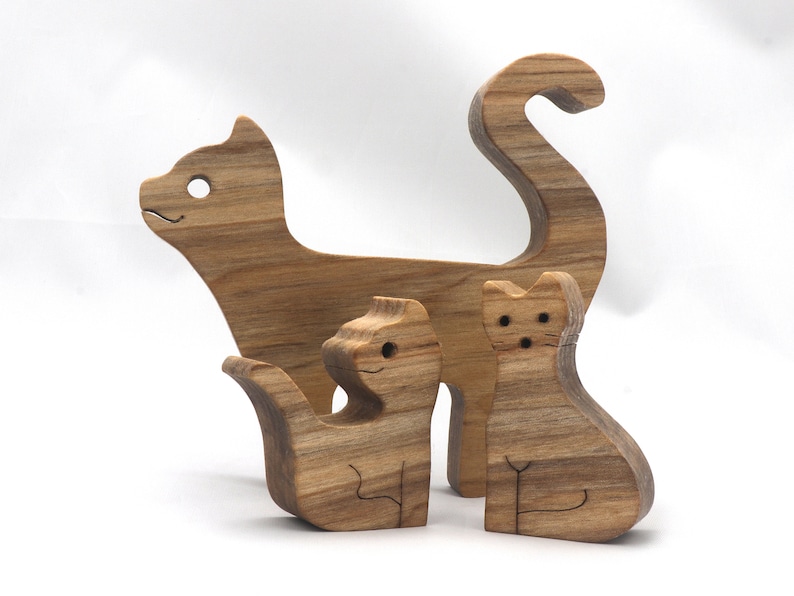 A freestanding wooden puzzle cut in the shape of a large cat standing on four legs. Two smaller kitten figures nest on the large cat’s back and interlock as puzzle pieces. Simple drilled holes form the eyes and nose on each cat, with a shallow cut line for the mouth on the large cat. The completed puzzle stands upright on a plain white surface. Handmade by Odin’s Toy Factory In Tallahassee, Florida, USA.