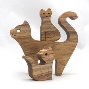 A freestanding wooden puzzle cut in the shape of a large cat standing on four legs. Two smaller kitten figures nest on the large cat’s back and interlock as puzzle pieces. Simple drilled holes form the eyes and nose on each cat, with a shallow cut line for the mouth on the large cat. The completed puzzle stands upright on a plain white surface. Handmade by Odin’s Toy Factory In Tallahassee, Florida, USA.
