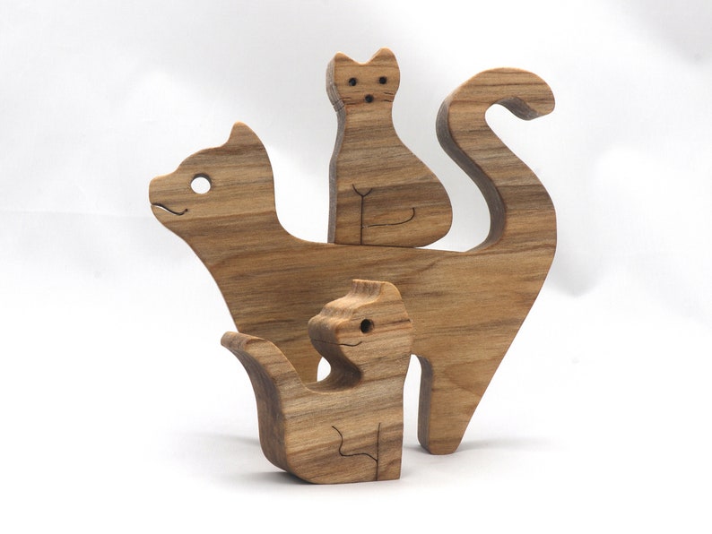 A freestanding wooden puzzle cut in the shape of a large cat standing on four legs. Two smaller kitten figures nest on the large cat’s back and interlock as puzzle pieces. Simple drilled holes form the eyes and nose on each cat, with a shallow cut line for the mouth on the large cat. The completed puzzle stands upright on a plain white surface. Handmade by Odin’s Toy Factory In Tallahassee, Florida, USA.