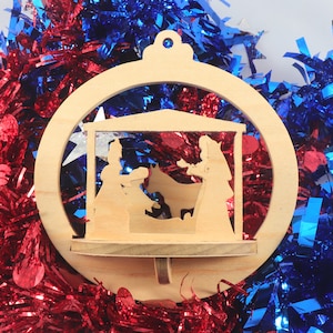 May include: A wooden Christmas ornament featuring a nativity scene within a circular frame. The ornament is set against a backdrop of red and blue tinsel, creating a festive and patriotic display. The ornament's design includes silhouettes of Mary, Joseph, and the baby Jesus.