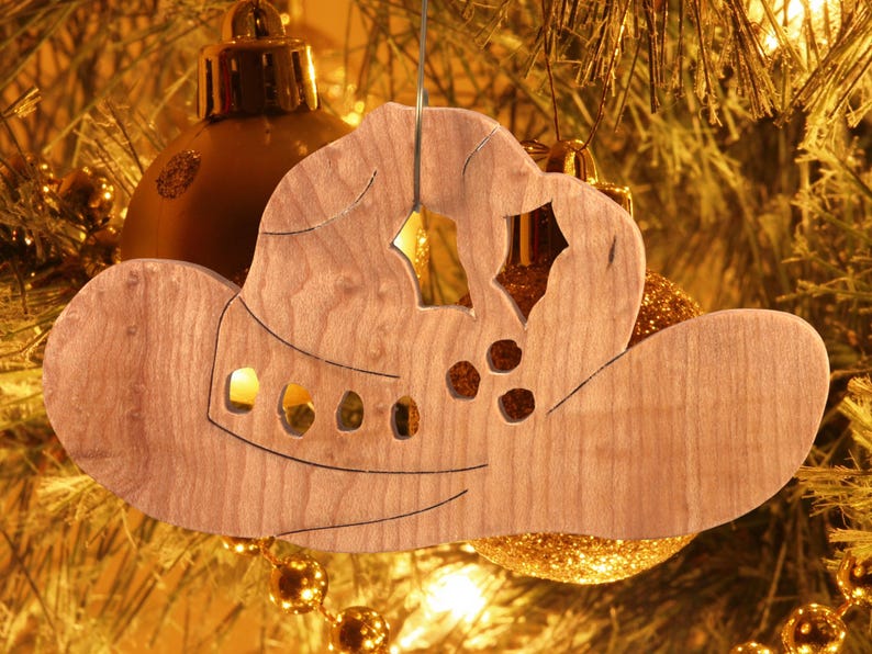 Handmade wooden Christmas tree ornament shaped like a cowboy hat, featuring a sprig of holly tucked into the hat band, star-shaped leaf cutouts, and round berry accents.