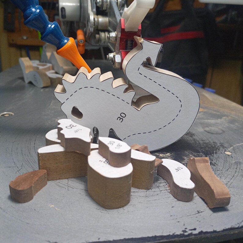 Unassembled wooden dragon parts on a scroll saw table. A paper cutting pattern covers the dragon’s body beside the saw blade, with smaller pieces spread in front.