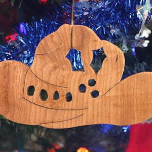 Handmade wooden Christmas tree ornament shaped like a cowboy hat, featuring a sprig of holly tucked into the hat band, star-shaped leaf cutouts, and round berry accents.