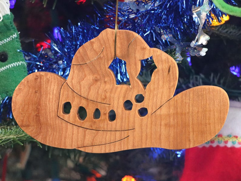 Handmade wooden Christmas tree ornament shaped like a cowboy hat, featuring a sprig of holly tucked into the hat band, star-shaped leaf cutouts, and round berry accents.