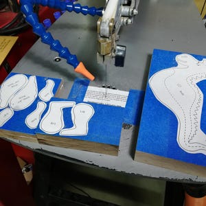 Baby dragon body, wing, leg, arm, and ear patterns attached to hardwood blanks with blue painter’s tape on a scroll saw.