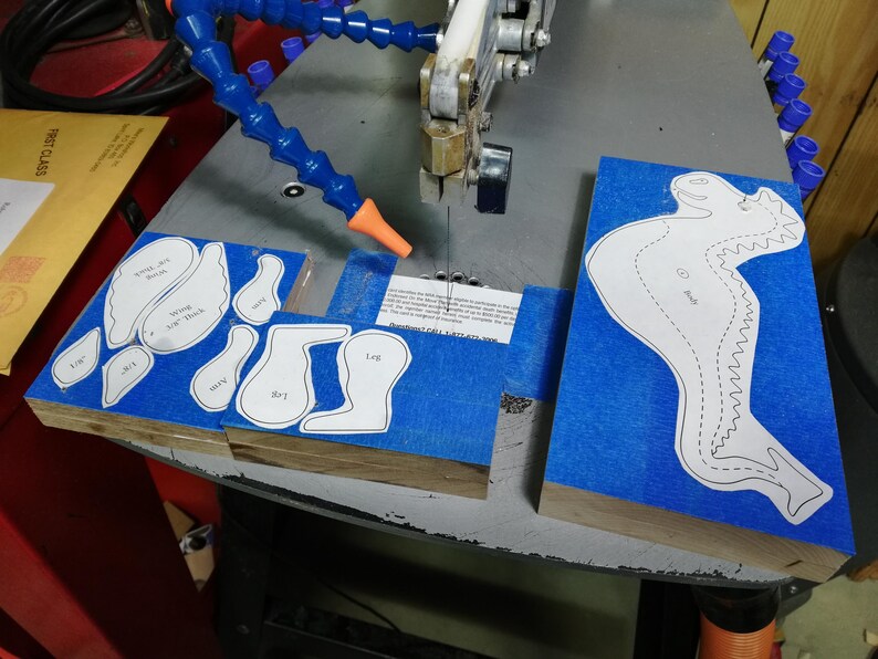 Baby dragon body, wing, leg, arm, and ear patterns attached to hardwood blanks with blue painter’s tape on a scroll saw.