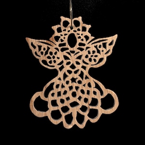 May include: A wooden angel ornament with intricate cut-out details. The angel has a halo, wings, and a flowing dress-like design. The ornament is made of light-colored wood and hangs from a thin silver string against a black background.