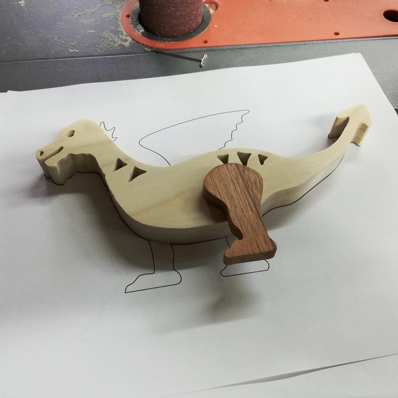 Partly assembled wooden dragon on a paper pattern in the workshop. The light-colored body has cutouts along its back, with one darker wooden leg laid against it and a sanding drum visible behind.