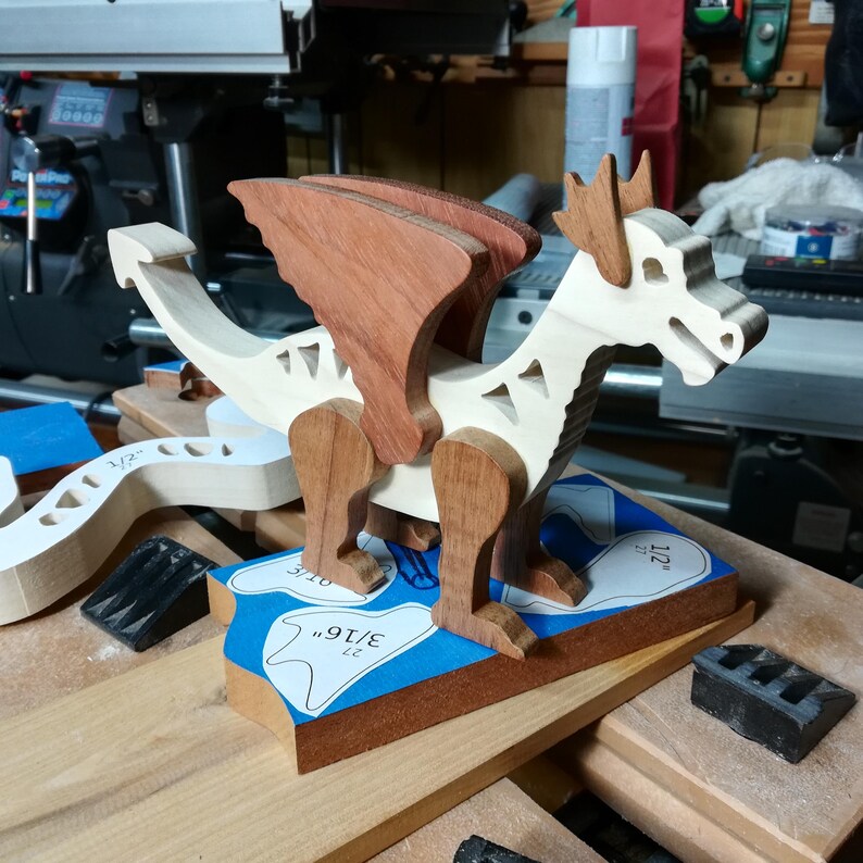 Wooden dragon figurine facing right on a workshop bench, showing its pale body and curved tail, brown wings, horns, and legs. Cut wooden pieces and paper patterns are nearby.