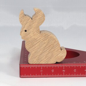 May include: A wooden rabbit figurine, painted in a light brown color, is sitting on a red measuring tool. The tool has markings in inches and fractions of an inch.