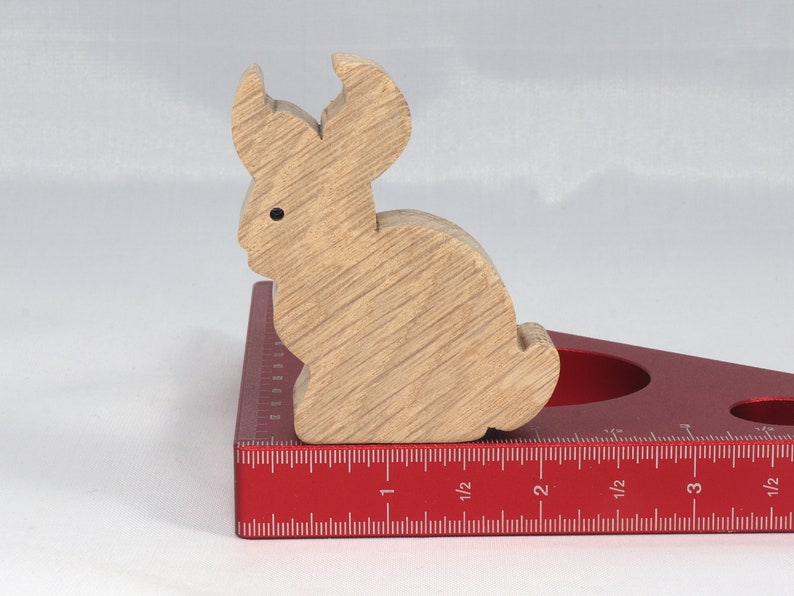 May include: A wooden rabbit figurine, painted in a light brown color, is sitting on a red measuring tool. The tool has markings in inches and fractions of an inch.