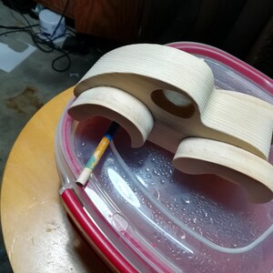 An unpainted wooden fat-fender toy car body rests on a clear plastic container lid with a red rim atop a round wooden stool. Viewed from above, the car has attached curved fenders, an oval window opening, and visible pine grain across the roof and hood. The wheels are absent, and a paint-marked wooden dowel projects from one axle hole. Workshop flooring, cords, and a paper offcut appear in the background.