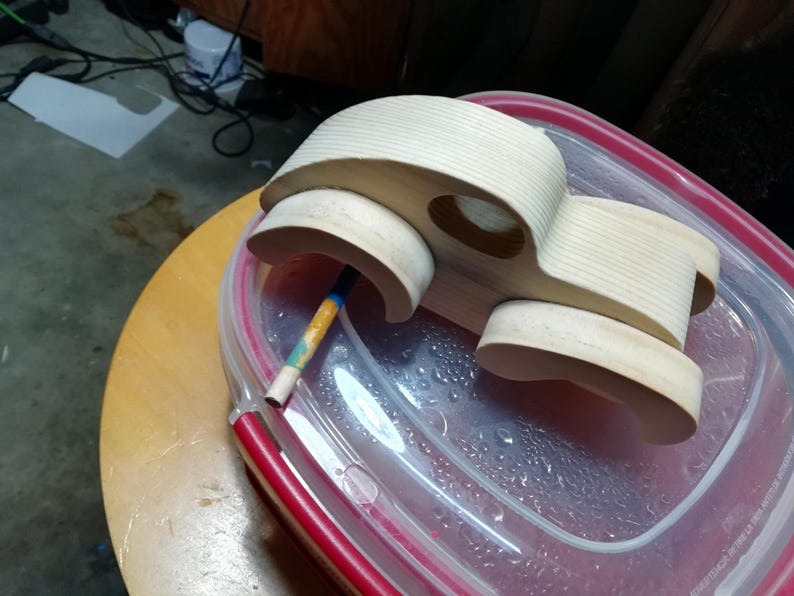 An unpainted wooden fat-fender toy car body rests on a clear plastic container lid with a red rim atop a round wooden stool. Viewed from above, the car has attached curved fenders, an oval window opening, and visible pine grain across the roof and hood. The wheels are absent, and a paint-marked wooden dowel projects from one axle hole. Workshop flooring, cords, and a paper offcut appear in the background.