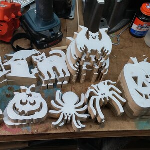 Handmade Halloween wood cutouts on a toymaker's workbench with paper patterns attached, including cats, bats, spiders, pumpkins, ghosts and skulls.