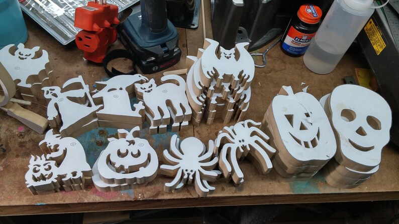 Handmade Halloween wood cutouts on a toymaker's workbench with paper patterns attached, including cats, bats, spiders, pumpkins, ghosts and skulls.