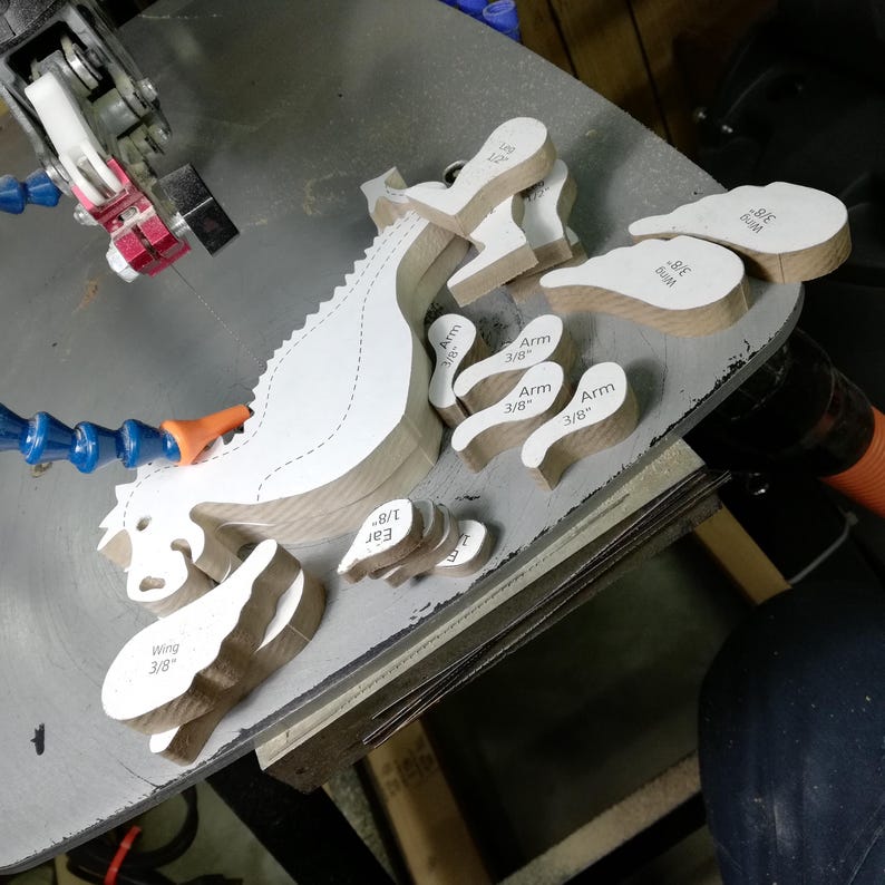 Freshly scroll-sawn wooden baby dragon body, wings, arms, legs, and ears arranged on the saw table before sanding and assembly.