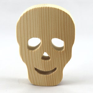 A handcrafted, unfinished light wood Halloween skull cutout featuring carefully carved eye sockets, a nose, and a cheerful, smiling mouth. Displayed upright against a white background.