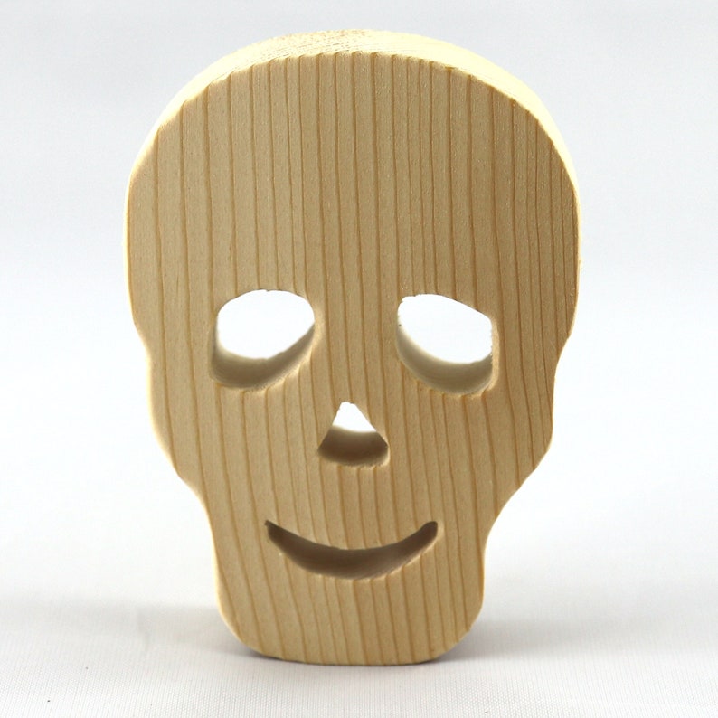 A handcrafted, unfinished light wood Halloween skull cutout featuring carefully carved eye sockets, a nose, and a cheerful, smiling mouth. Displayed upright against a white background.