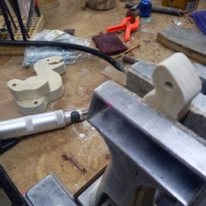 Unfinished wooden dinosaur toy parts on a workbench, with a drilled wooden part held upright in a bench vise among sanding supplies and tools.