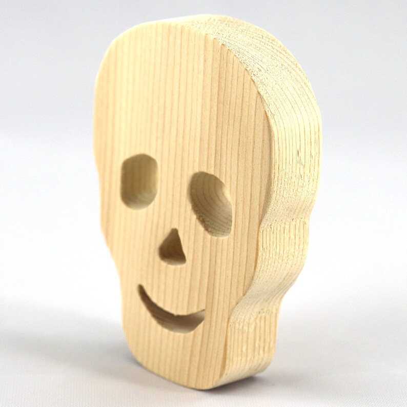 A handcrafted, unfinished light wood Halloween skull cutout featuring carefully carved eye sockets, a nose, and a cheerful, smiling mouth. Displayed upright against a white background.