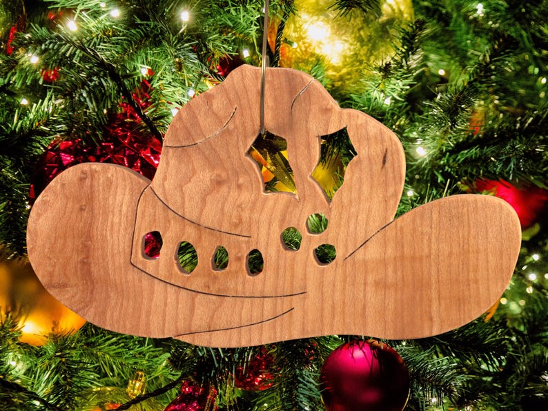 Handmade wooden Christmas tree ornament shaped like a cowboy hat, featuring a sprig of holly tucked into the hat band, star-shaped leaf cutouts, and round berry accents.