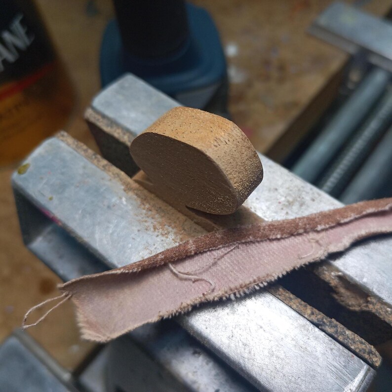 Close-up of a small dark wood dragon part held in a metal vise, with a strip of sandpaper laid across the work area.
