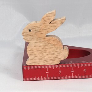 May include: A wooden rabbit figurine, painted in a light brown color, is sitting on a red measuring tool. The tool has a ruler marked in inches, with markings for 1, 1 1/2, 2, 2 1/2, and 3 inches.