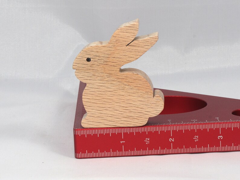May include: A wooden rabbit figurine, painted in a light brown color, is sitting on a red measuring tool. The tool has a ruler marked in inches, with markings for 1, 1 1/2, 2, 2 1/2, and 3 inches.