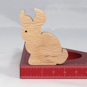 May include: A wooden rabbit figurine, painted in a light brown color, is sitting on a red measuring tool with a ruler marked in inches. The rabbit is about 2 inches tall.