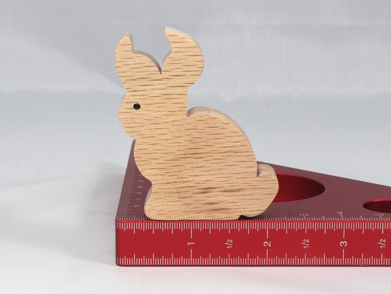 May include: A wooden rabbit figurine, painted in a light brown color, is sitting on a red measuring tool with a ruler marked in inches. The rabbit is about 2 inches tall.