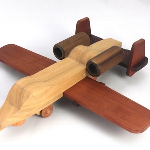 May include: A wooden toy airplane with a light brown fuselage, dark brown engine housings, and reddish-brown wings and tail. The toy has small wooden wheels and is designed to resemble a military aircraft.