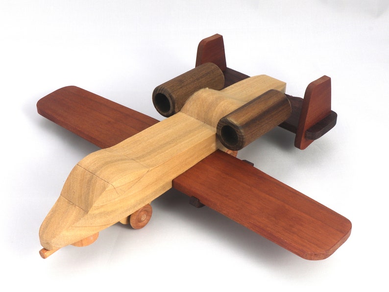 May include: A wooden toy airplane with a light brown fuselage, dark brown engine housings, and reddish-brown wings and tail. The toy has small wooden wheels and is designed to resemble a military aircraft.