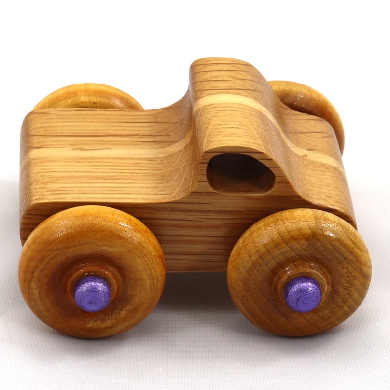 May include: A wooden toy truck with a natural wood grain finish. The truck has four large wooden wheels with purple center accents. The body of the truck is carved with a cab and a small opening. A classic toy for children.