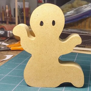 A handmade, unpainted wooden ghost cutout stands upright on a white background. The light-colored solid wood figure has a rounded head with two circular eye cutouts, raised wavy arms, and a curved wavy base, ready for painting or crafting. On my workbench.