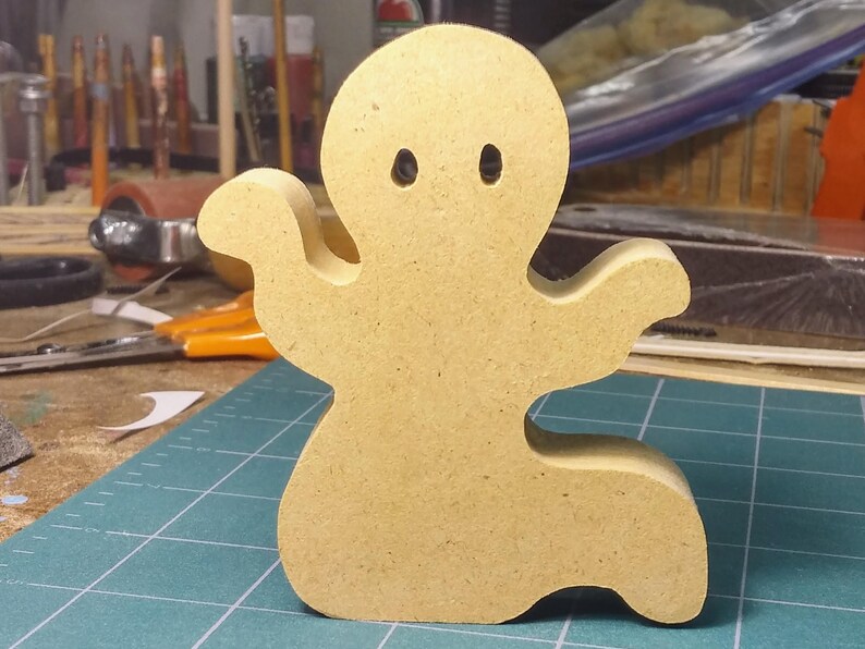 A handmade, unpainted wooden ghost cutout stands upright on a white background. The light-colored solid wood figure has a rounded head with two circular eye cutouts, raised wavy arms, and a curved wavy base, ready for painting or crafting. On my workbench.