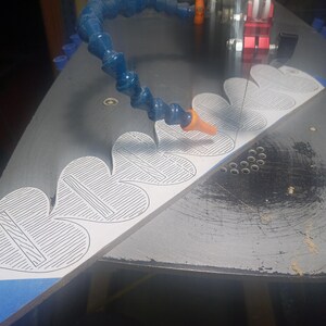 May include: A white wooden board with a series of heart-shaped outlines featuring internal line patterns, positioned on a black scroll saw table. A blue flexible coolant hose is directed toward the blade, which is cutting the wood material.