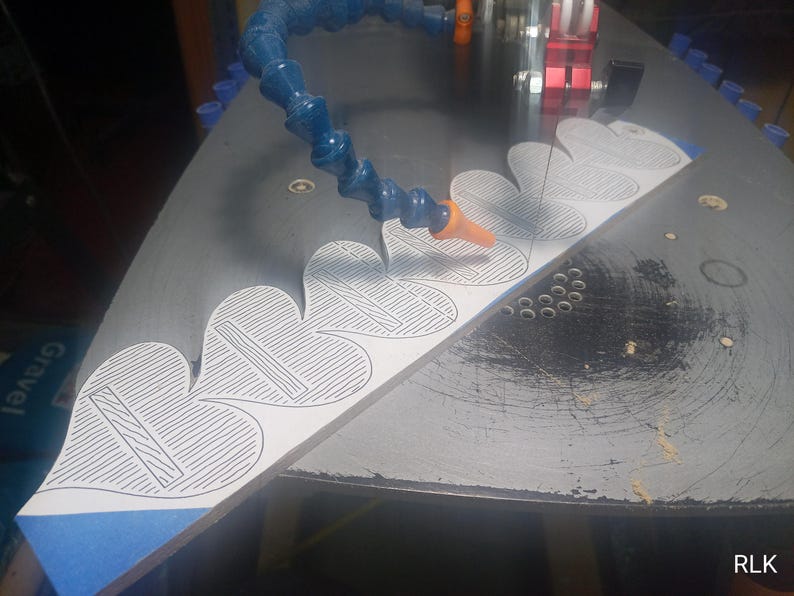 May include: A white wooden board with a series of heart-shaped outlines featuring internal line patterns, positioned on a black scroll saw table. A blue flexible coolant hose is directed toward the blade, which is cutting the wood material.