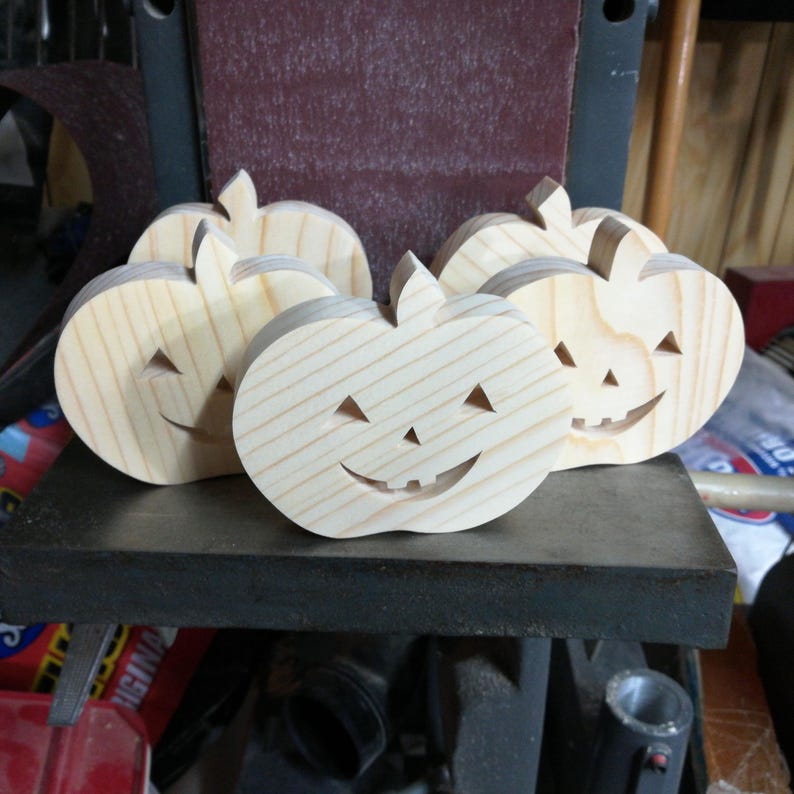 May include: A set of four light-colored wooden pumpkin cutouts featuring carved triangular eyes, a small triangular nose, and a wide smiling mouth with visible teeth. Each pumpkin has a small stem at the top and a natural wood grain finish.