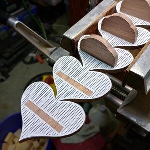 May include: A series of heart-shaped wooden objects held in a metal vise. Each heart features a white surface with thin black vertical stripes and a horizontal rectangular wooden inlay. A small wooden arch is attached to the top of each heart shape.