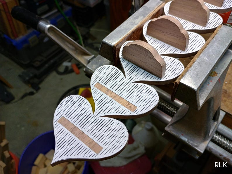 May include: A series of heart-shaped wooden objects held in a metal vise. Each heart features a white surface with thin black vertical stripes and a horizontal rectangular wooden inlay. A small wooden arch is attached to the top of each heart shape.