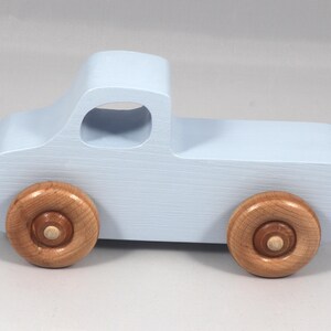May include: A light blue wooden toy truck with natural wood wheels. The truck has a classic pickup design with a cab and a cargo bed. The wheels are round and attached to the truck's body.