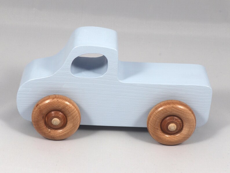 May include: A light blue wooden toy truck with natural wood wheels. The truck has a classic pickup design with a cab and a cargo bed. The wheels are round and attached to the truck's body.