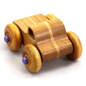 May include: A handcrafted wooden toy car with a natural wood finish. The car features four large, round wheels with purple center accents. The body of the car is made of light brown wood with a curved design.