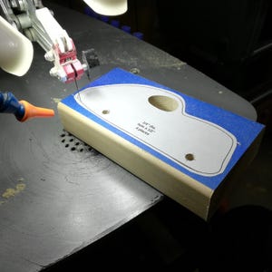 A pine block rests on a scroll saw table as the blade follows the curved outline of a wooden toy car body. A paper pattern attached over blue painter’s tape marks the fat-fender car’s profile. The oval window opening and two axle holes have already been cut or drilled. An orange air nozzle points toward the cutting area, with sawdust scattered across the metal scroll saw table.