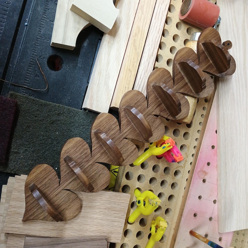 May include: A series of five connected wooden heart-shaped wall hooks arranged in a diagonal line. Each heart features a dark brown wood grain finish and a small horizontal shelf or hook protruding from the center of the heart shape.