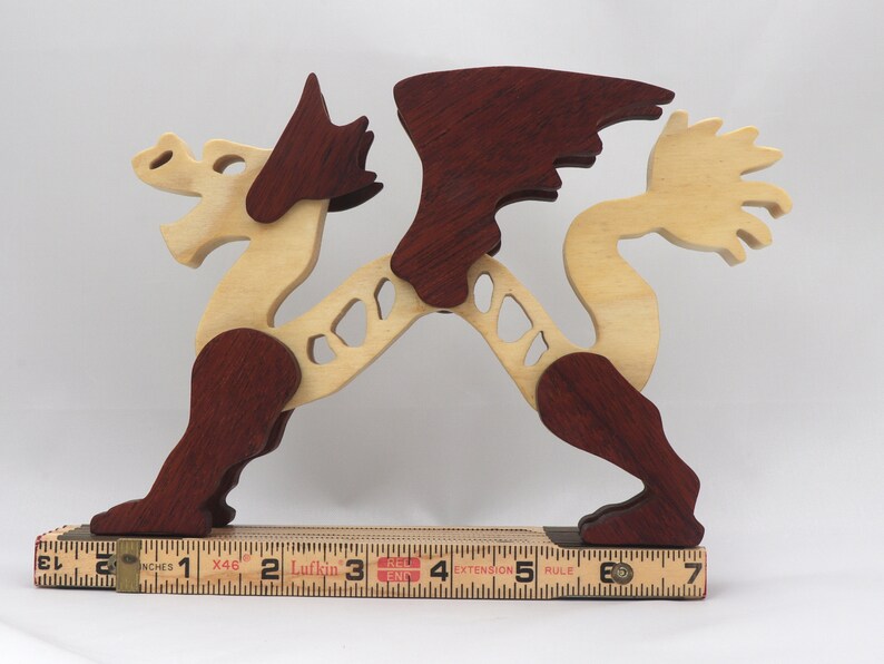 Handmade wooden dragon figurine. crafted from contrasting hardwoods with a light natural wood body and head paired with rich dark reddish-brown wings, legs, and horns. Displayed on a folding carpenters rule for scale.