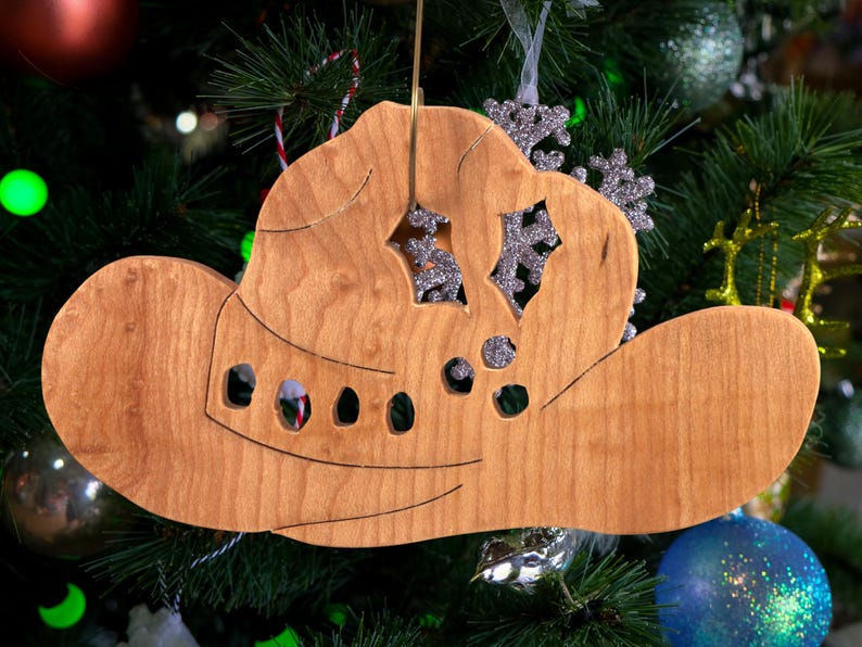 Handmade wooden Christmas tree ornament shaped like a cowboy hat, featuring a sprig of holly tucked into the hat band, star-shaped leaf cutouts, and round berry accents.