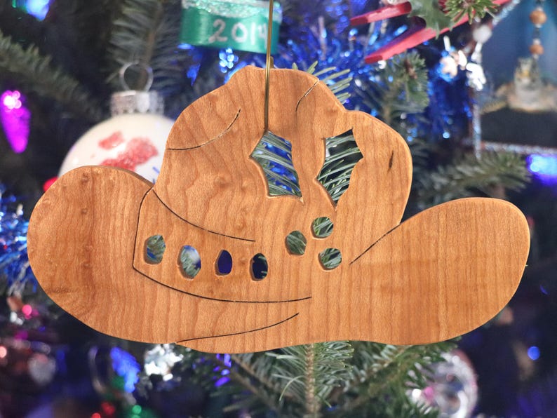 Handmade wooden Christmas tree ornament shaped like a cowboy hat, featuring a sprig of holly tucked into the hat band, star-shaped leaf cutouts, and round berry accents.