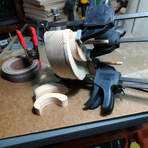An unfinished pine toy car body stands on its side on a workshop bench while black bar clamps hold two curved fat fenders against it. A positioning template aligns the fenders, and wooden dowels project from the axle holes to align the template. Two loose, sanded fenders rest in the foreground. A rolled sanding belt secured with a red-handled spring clamp sits to the left, with tools and stacked wood in the background.